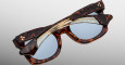 The Jacques Marie Mage Small Elmyr tortoiseshell sunglasses with blue lenses are folded closed on a plain light background, showcasing two star-like details on the temple hinges.
