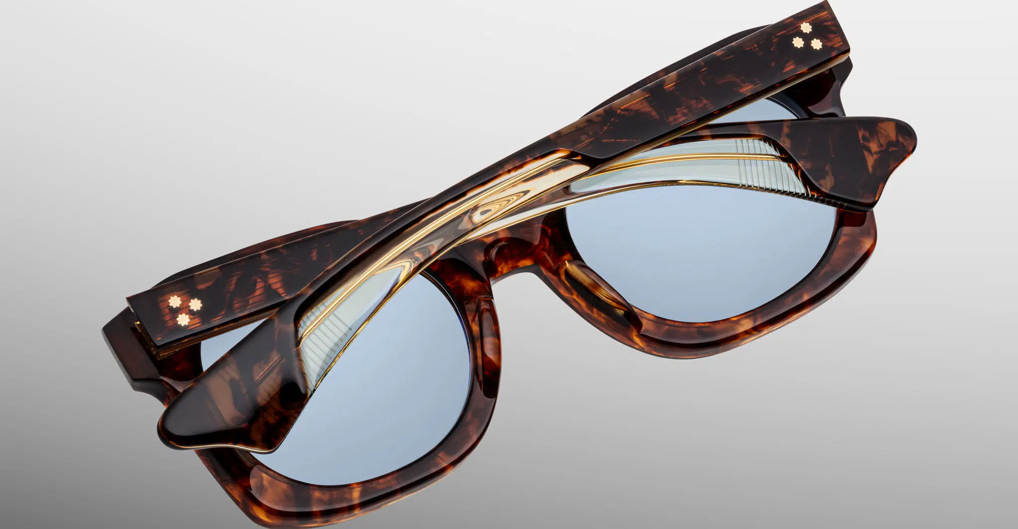 The Jacques Marie Mage Small Elmyr tortoiseshell sunglasses with blue lenses are folded closed on a plain light background, showcasing two star-like details on the temple hinges.