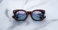 The Jacques Marie Mage Small Elmyr tortoiseshell sunglasses with blue-tinted lenses rest on white marble, their reflective lenses capturing the image of a modern bridge with railings.