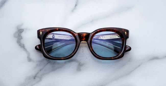 The Jacques Marie Mage Small Elmyr tortoiseshell sunglasses with blue-tinted lenses rest on white marble, their reflective lenses capturing the image of a modern bridge with railings.