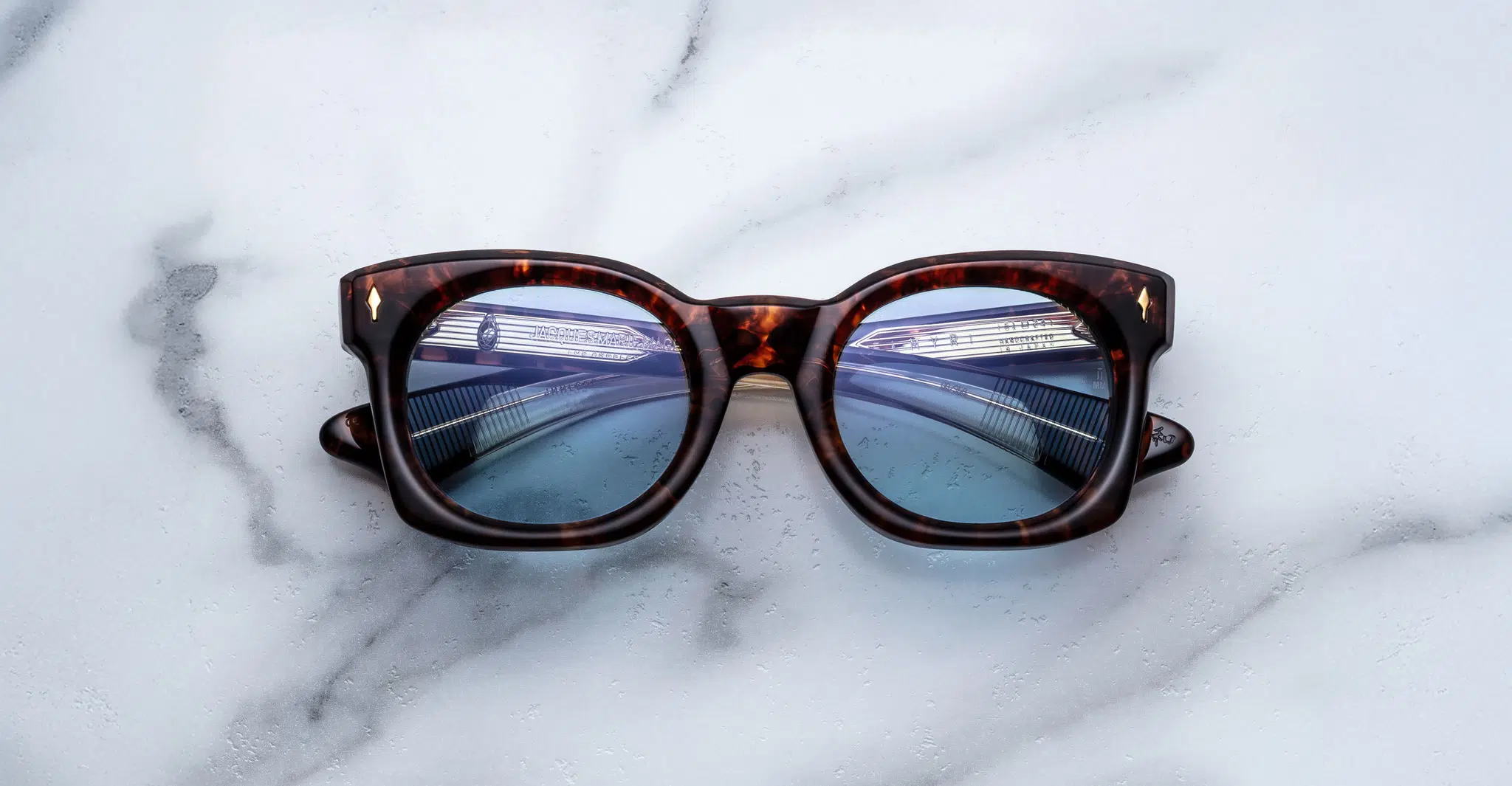 The Jacques Marie Mage Small Elmyr tortoiseshell sunglasses with blue-tinted lenses rest on white marble, their reflective lenses capturing the image of a modern bridge with railings.