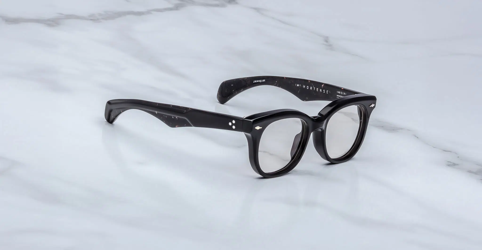 The Jacques Marie Mage Hortense eyeglasses feature black, thick round frames with subtle white speckles on the arms and silver accents by the hinges, displayed on a white marble surface.