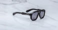 The Jacques Marie Mage Jasper sunglasses, featuring black thick frames, dark lenses, and silver dot accents near the hinges, rest on a white marble surface.