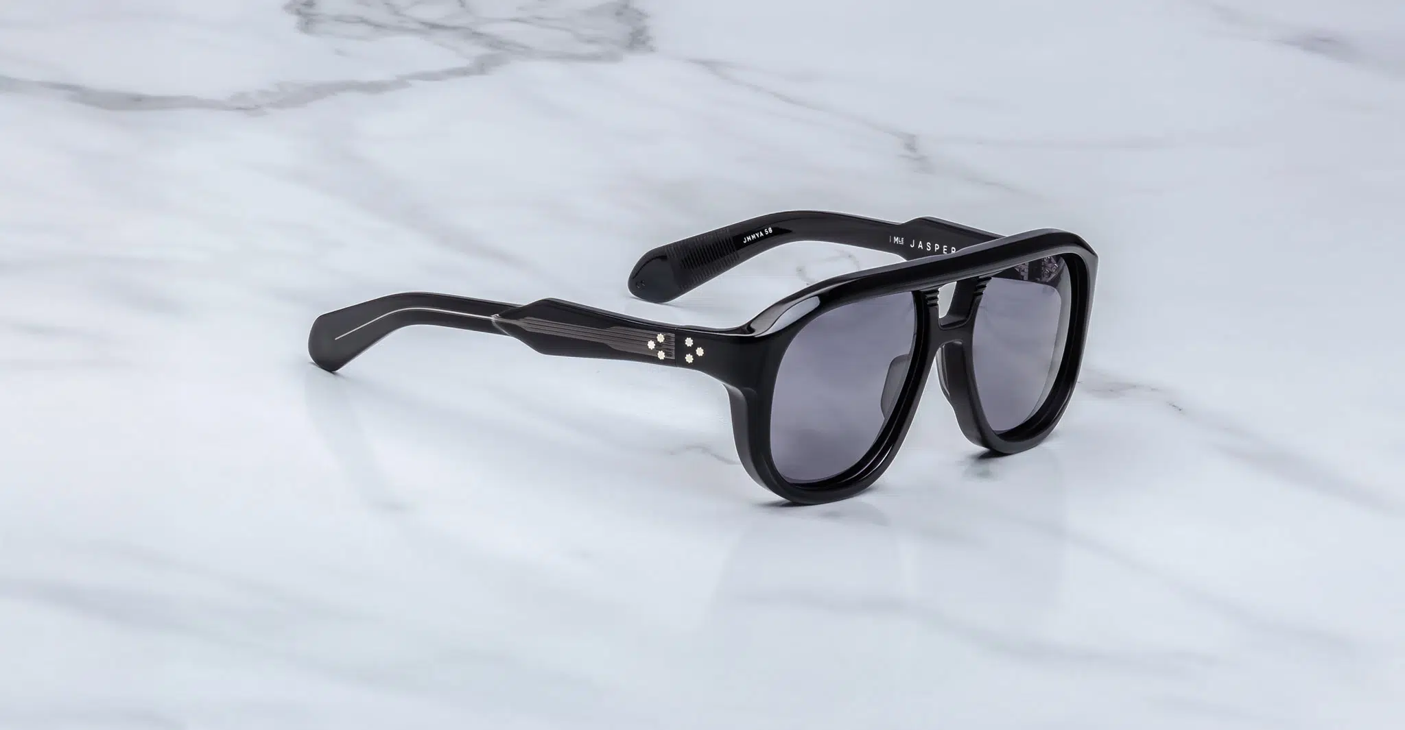 The Jacques Marie Mage Jasper sunglasses, featuring black thick frames, dark lenses, and silver dot accents near the hinges, rest on a white marble surface.