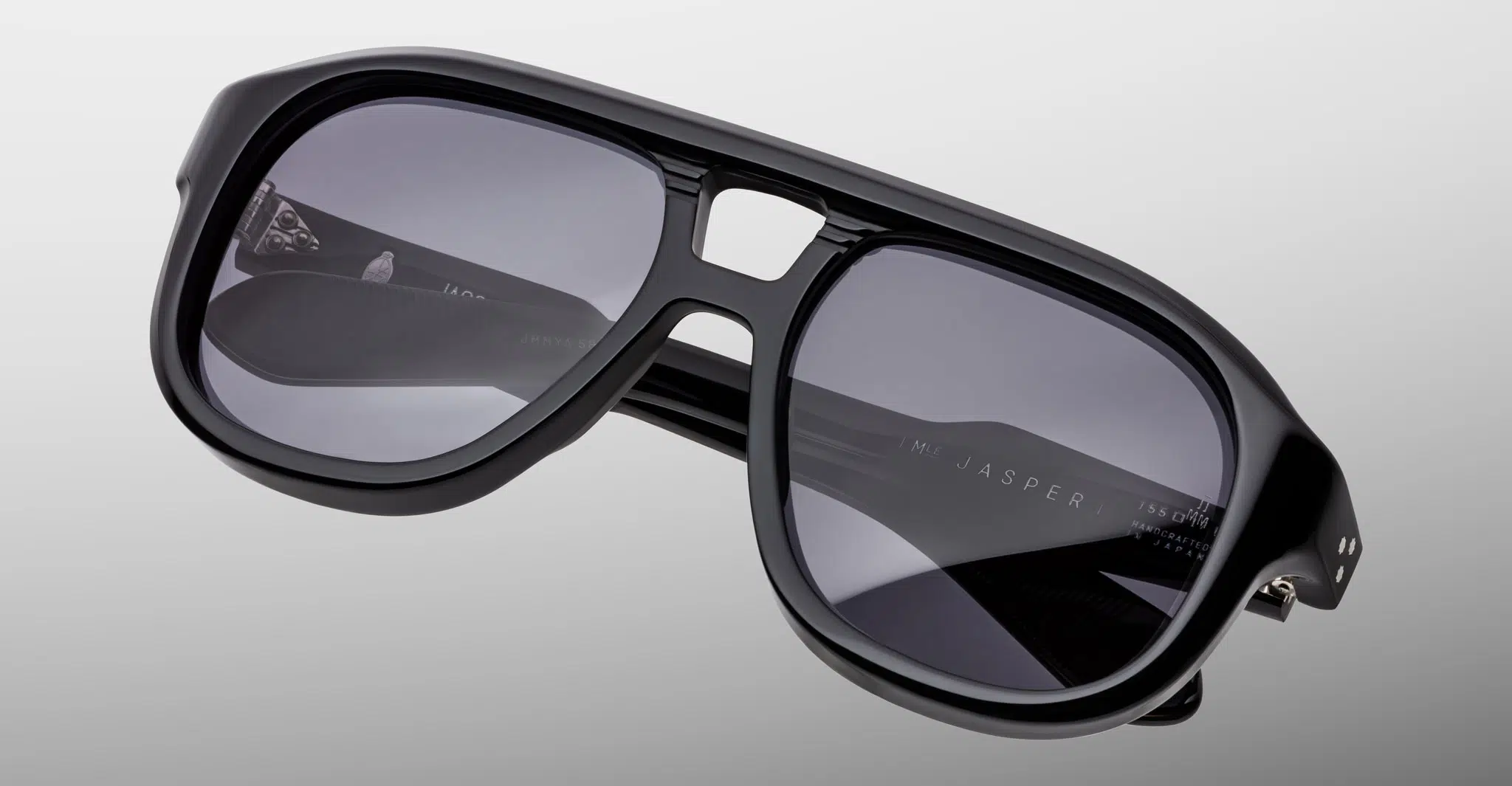 The Jacques Marie Mage Jasper sunglasses feature an oversized black frame with dark tinted lenses, a bold thick design, and a double bridge, displayed on a gradient gray background.