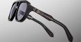 Close-up of Jacques Marie Mage Jasper black sunglasses with thick frames and dark lenses, featuring metallic circuit-like designs on the temples, set against a plain gradient gray background.