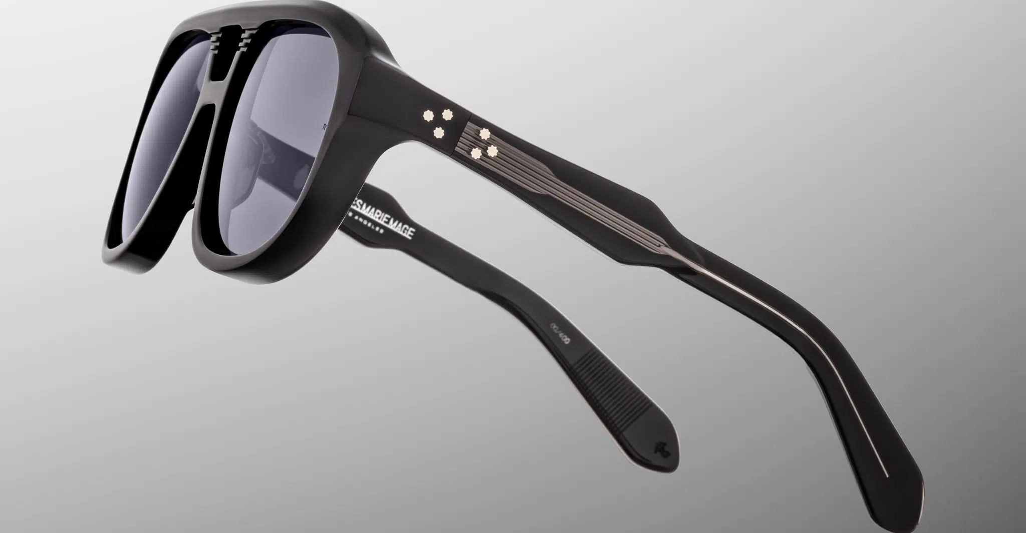 Close-up of Jacques Marie Mage Jasper black sunglasses with thick frames and dark lenses, featuring metallic circuit-like designs on the temples, set against a plain gradient gray background.