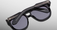 The Jacques Marie Mage Jasper sunglasses feature stylish black thick frames and dark lenses, shown folded closed and resting on a reflective gray surface.