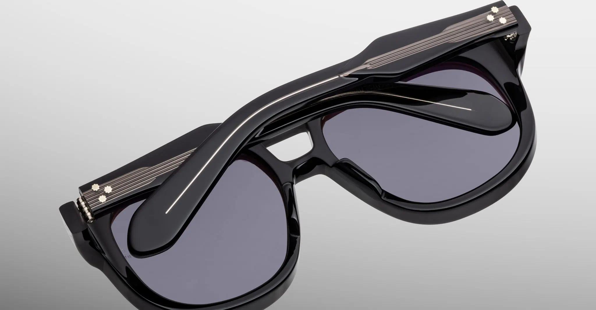 The Jacques Marie Mage Jasper sunglasses feature stylish black thick frames and dark lenses, shown folded closed and resting on a reflective gray surface.