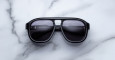 The Jacques Marie Mage Jasper sunglasses, featuring a thick black frame, dark lenses, and a metallic bridge accent, rest on a white marble surface.