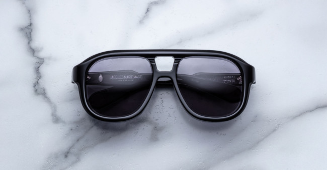The Jacques Marie Mage Jasper sunglasses, featuring a thick black frame, dark lenses, and a metallic bridge accent, rest on a white marble surface.
