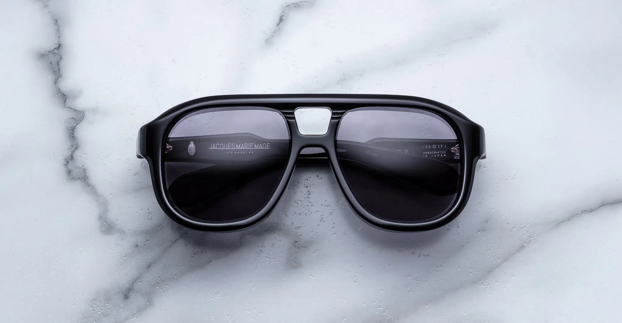 The Jacques Marie Mage Jasper sunglasses, featuring a thick black frame, dark lenses, and a metallic bridge accent, rest on a white marble surface.