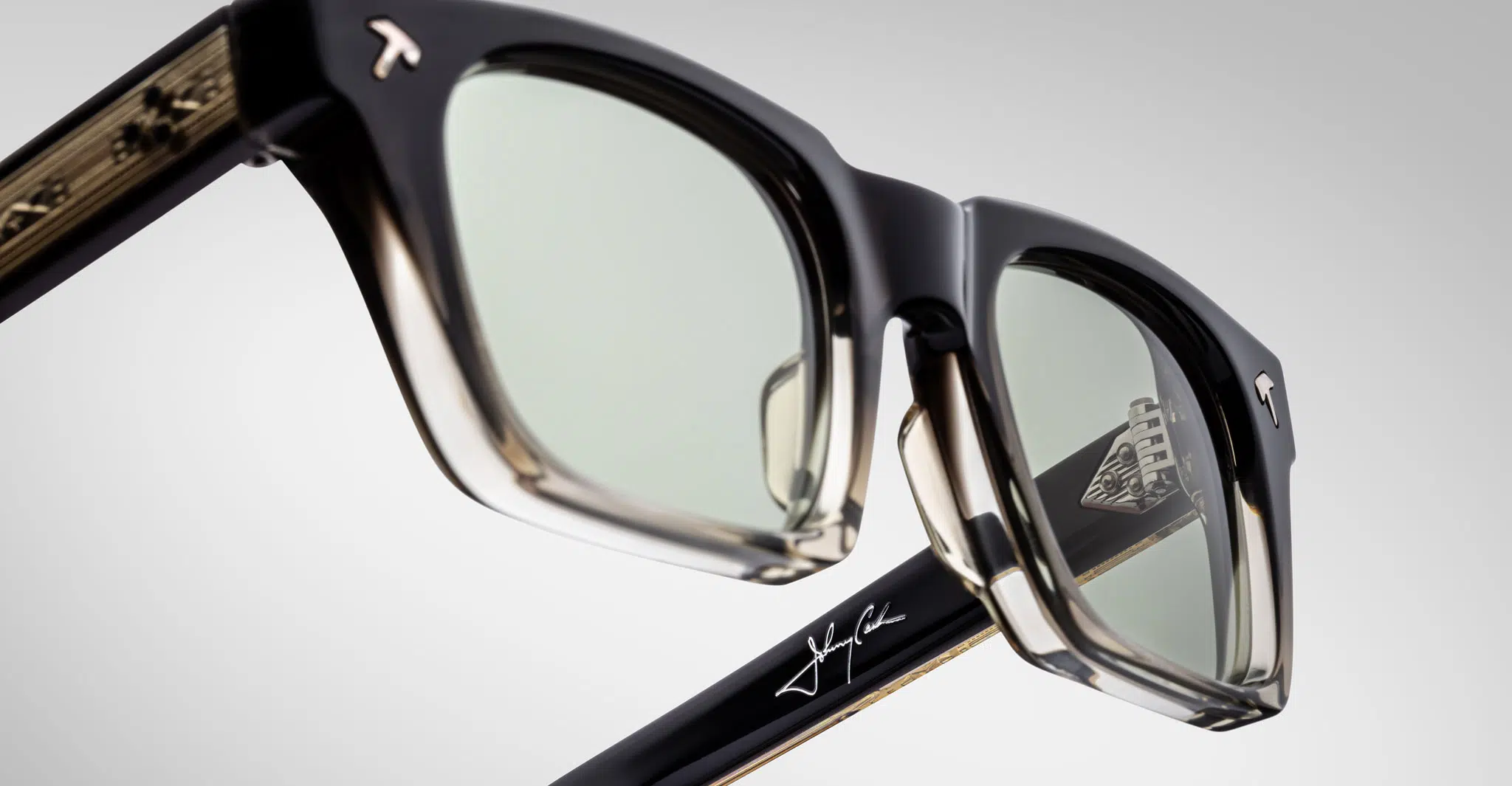 Close-up of Jacques Marie Mage Quentin sunglasses in black and translucent acetate, with tinted lenses, a Tesla “T” logo on the frame, engraved signature on the arm, and partial geometric pattern inside.