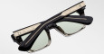 The Jacques Marie Mage Quentin sunglasses are shown folded, featuring black frames with gold detailing on the arms and top, and green-tinted lenses set against a plain light background.