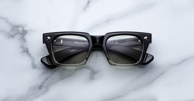 The Jacques Marie Mage Quentin eyeglasses feature bold black square frames with thick rims and metallic arrow accents on the corners, displayed on a white marble surface with lenses reflecting text and lines.