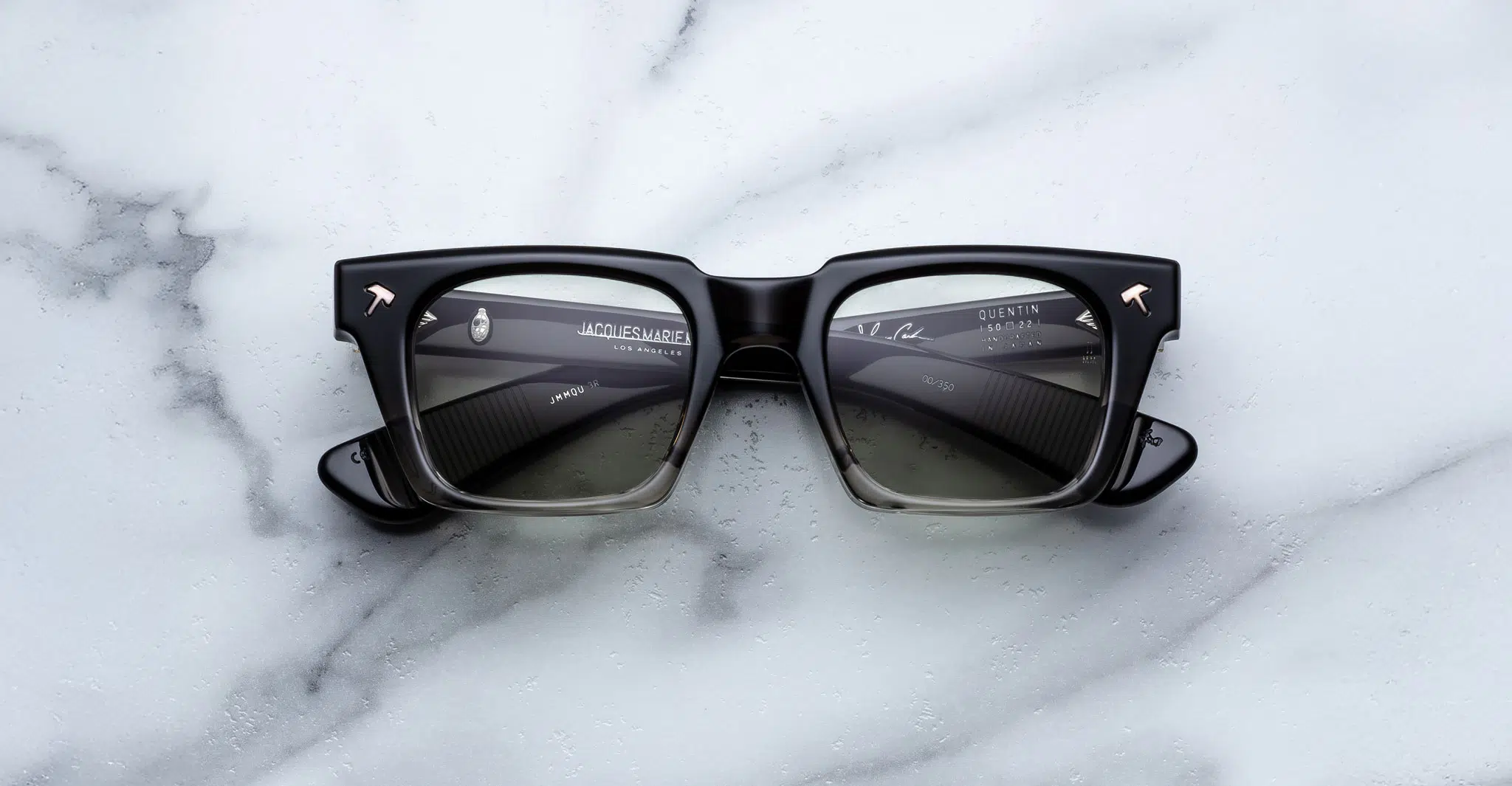 The Jacques Marie Mage Quentin eyeglasses feature bold black square frames with thick rims and metallic arrow accents on the corners, displayed on a white marble surface with lenses reflecting text and lines.