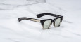 The Jacques Marie Mage Quentin sunglasses feature a black and translucent square frame with green-tinted lenses, thick arms adorned with metallic accents, and visible branding, shown resting on a white marble surface.