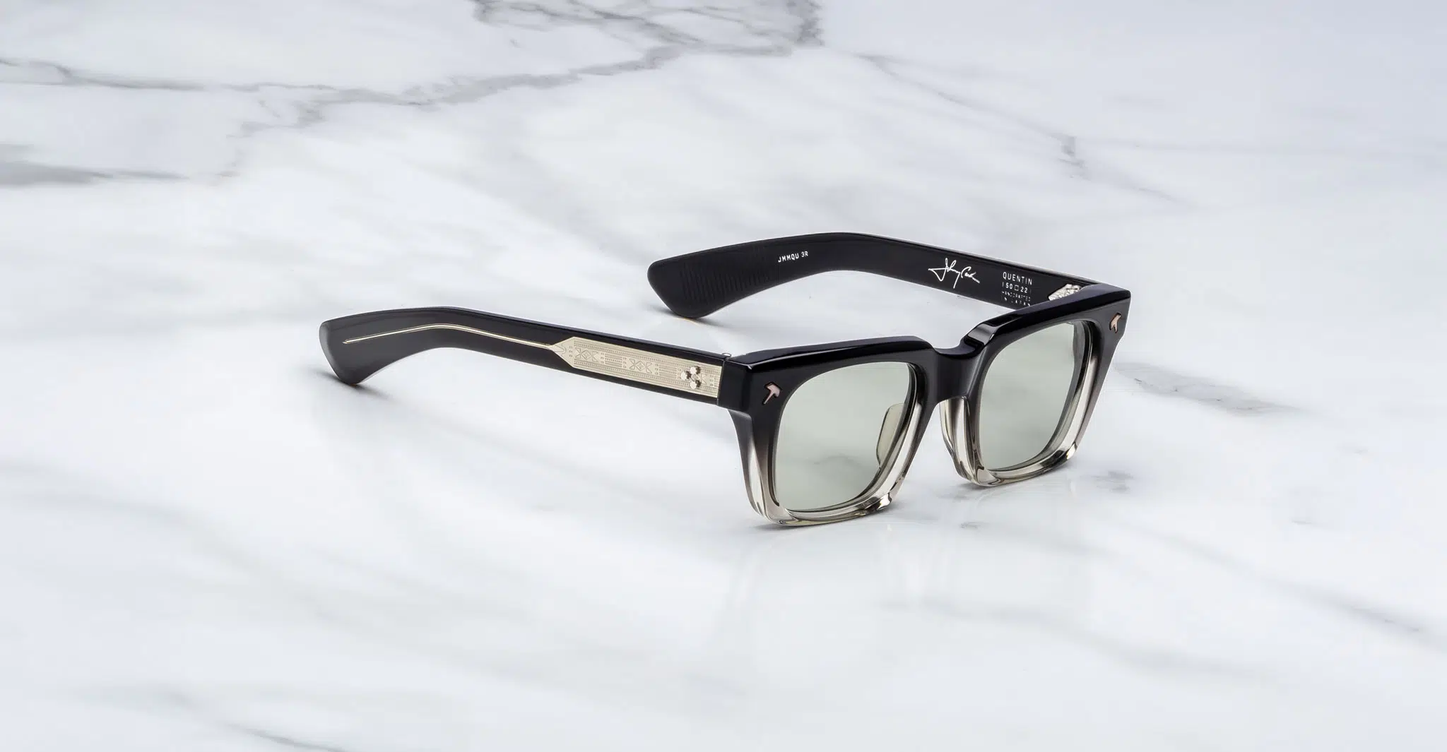 The Jacques Marie Mage Quentin sunglasses feature a black and translucent square frame with green-tinted lenses, thick arms adorned with metallic accents, and visible branding, shown resting on a white marble surface.