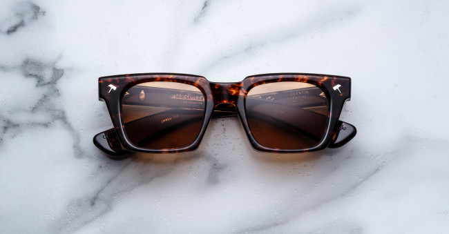 A pair of Jacques Marie Mage Quentin rectangular tortoiseshell sunglasses with brown lenses rests on a white marble surface, featuring subtle reflections and small logo markings on the lenses and frame.