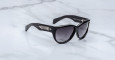 The Jacques Marie Mage Ravello sunglasses, featuring dark lenses, thick frames, and temples adorned with silver metal diamond-shaped detailing, are displayed on a white marble surface.