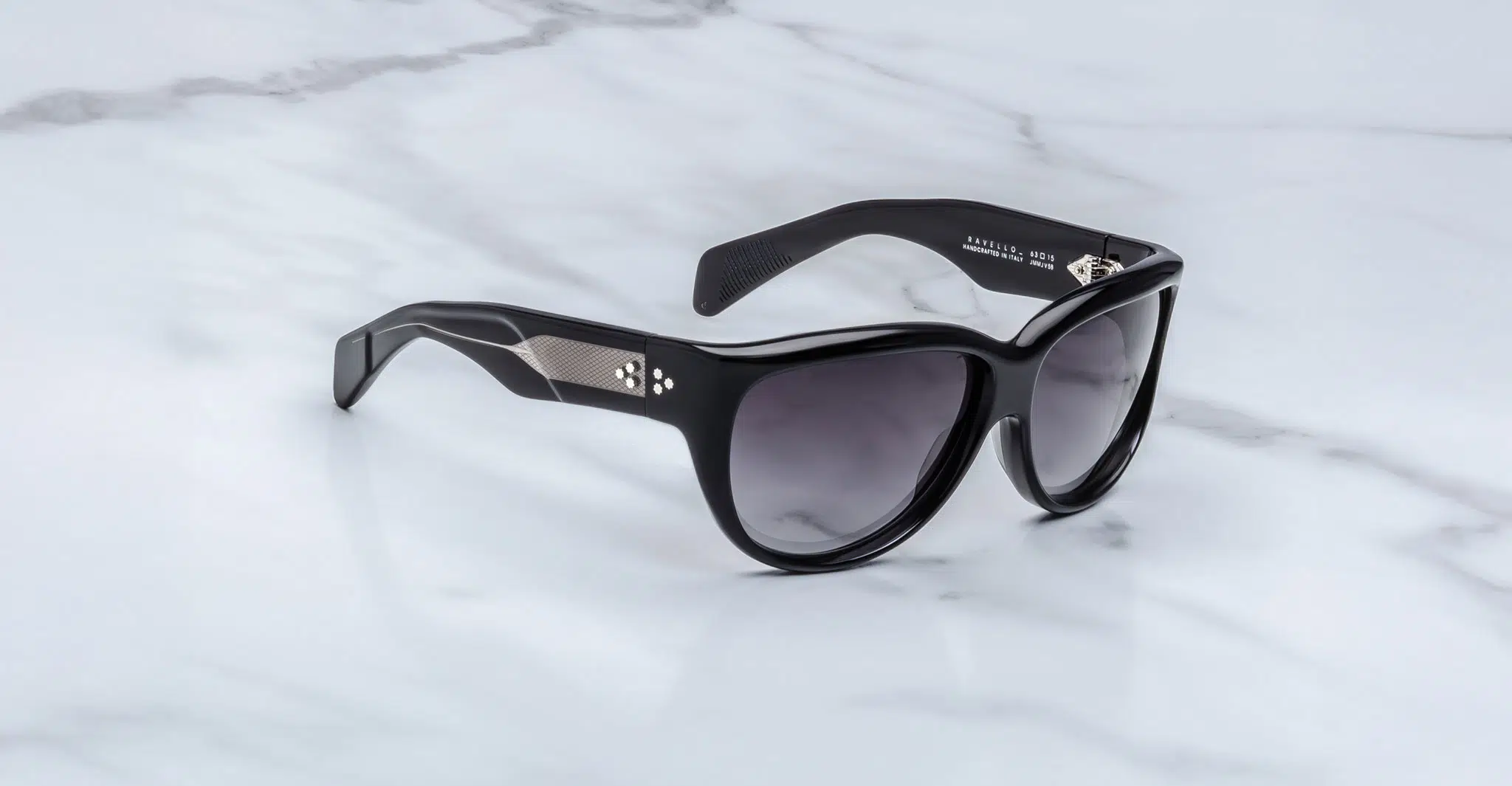 The Jacques Marie Mage Ravello sunglasses, featuring dark lenses, thick frames, and temples adorned with silver metal diamond-shaped detailing, are displayed on a white marble surface.