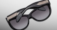 The Jacques Marie Mage Ravello sunglasses feature black frames, dark gradient lenses, metallic gold accents, and layered sculptural temples, shown against a plain gray background.