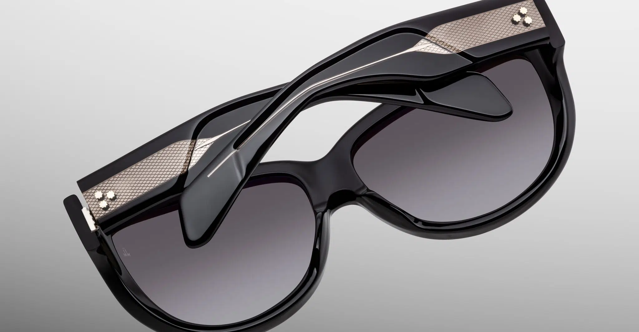 The Jacques Marie Mage Ravello sunglasses feature black frames, dark gradient lenses, metallic gold accents, and layered sculptural temples, shown against a plain gray background.