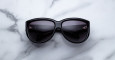 A Jacques Marie Mage Ravello pair of black sunglasses with dark lenses is placed on a white marble surface with grey veining, the lenses facing upward.
