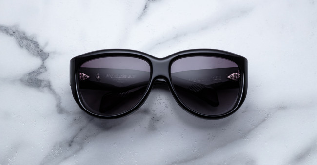 A Jacques Marie Mage Ravello pair of black sunglasses with dark lenses is placed on a white marble surface with grey veining, the lenses facing upward.