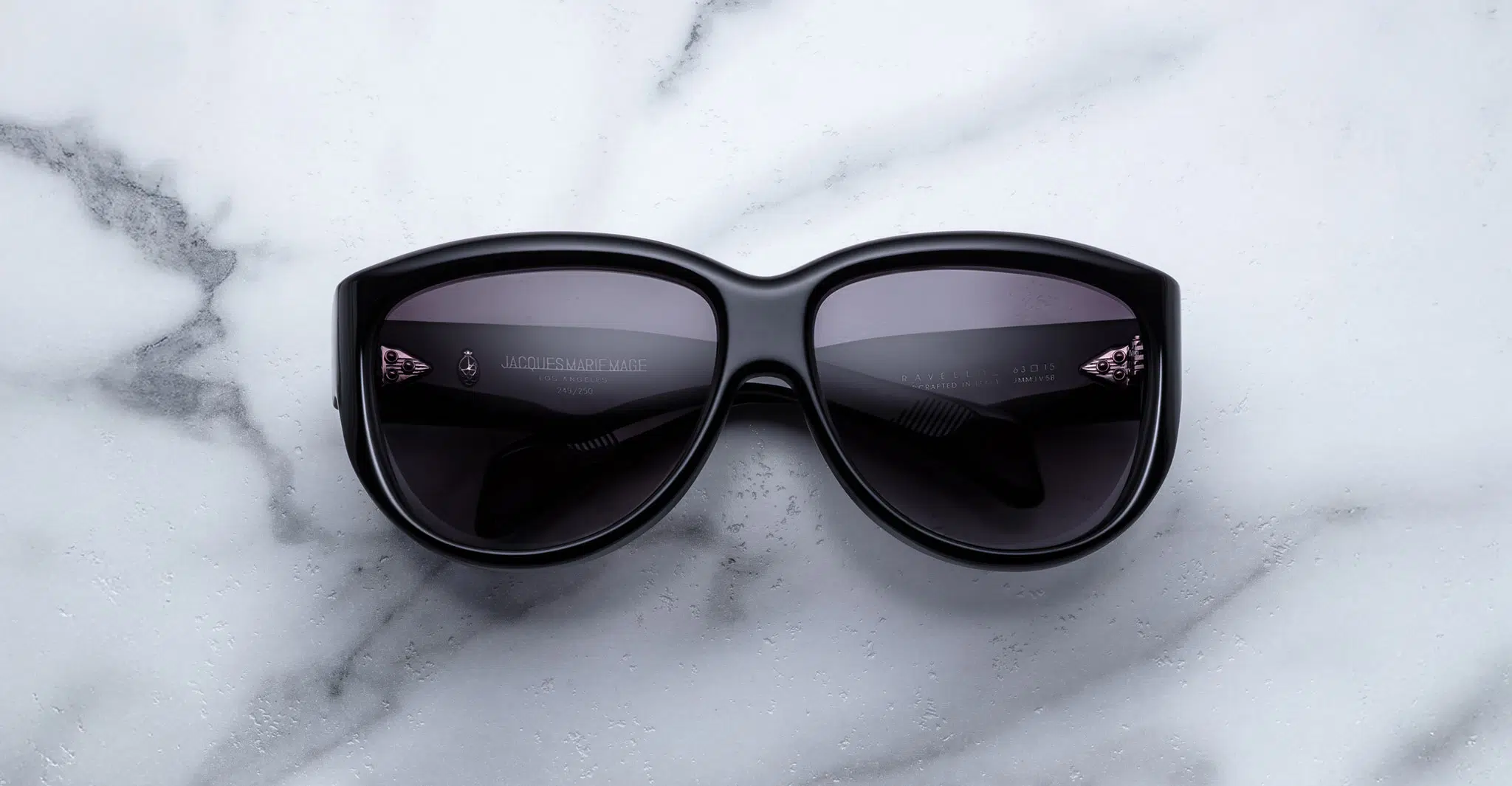 A Jacques Marie Mage Ravello pair of black sunglasses with dark lenses is placed on a white marble surface with grey veining, the lenses facing upward.