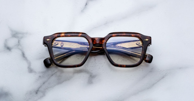 A pair of Jacques Marie Mage Tomlin tortoiseshell rectangular eyeglasses rests on a white marble surface, with the lenses reflecting a metallic object featuring horizontal lines.