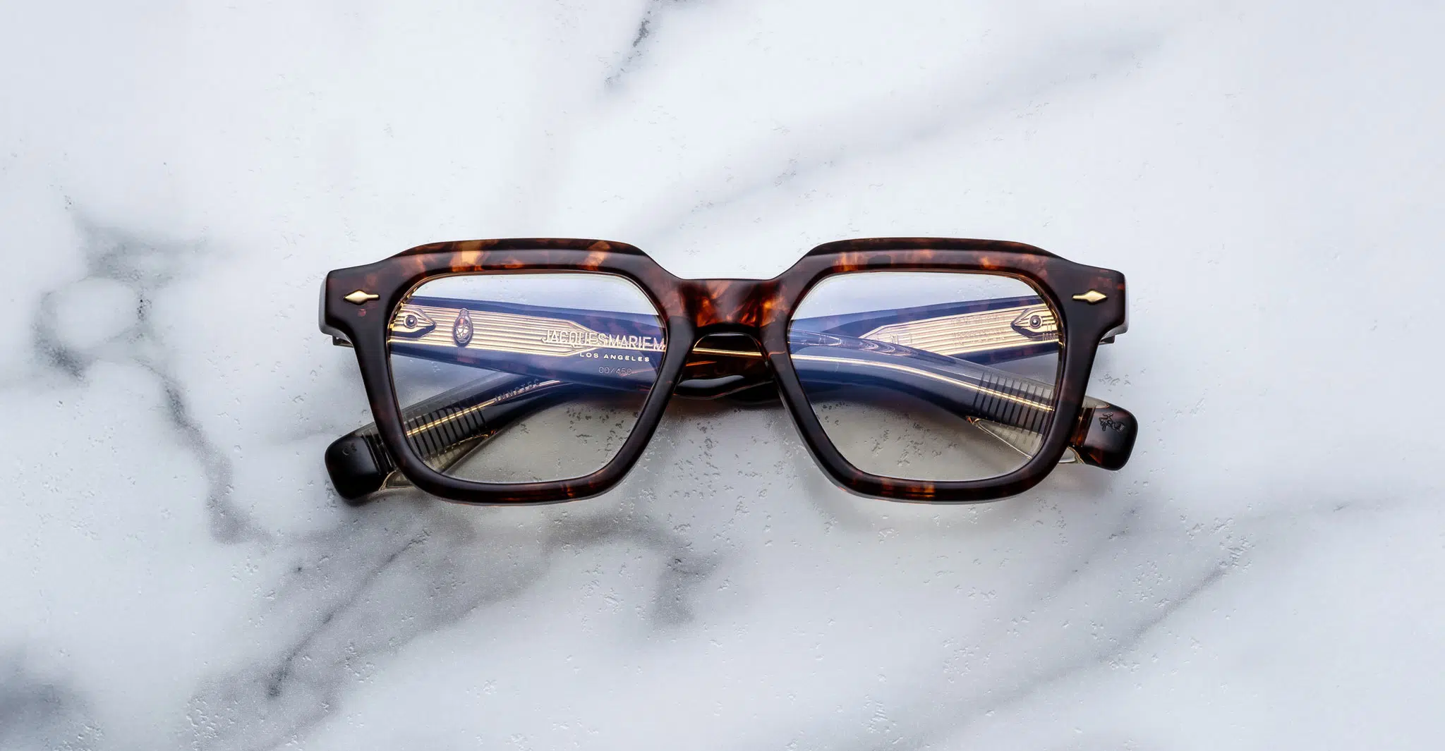 A pair of Jacques Marie Mage Tomlin tortoiseshell rectangular eyeglasses rests on a white marble surface, with the lenses reflecting a metallic object featuring horizontal lines.