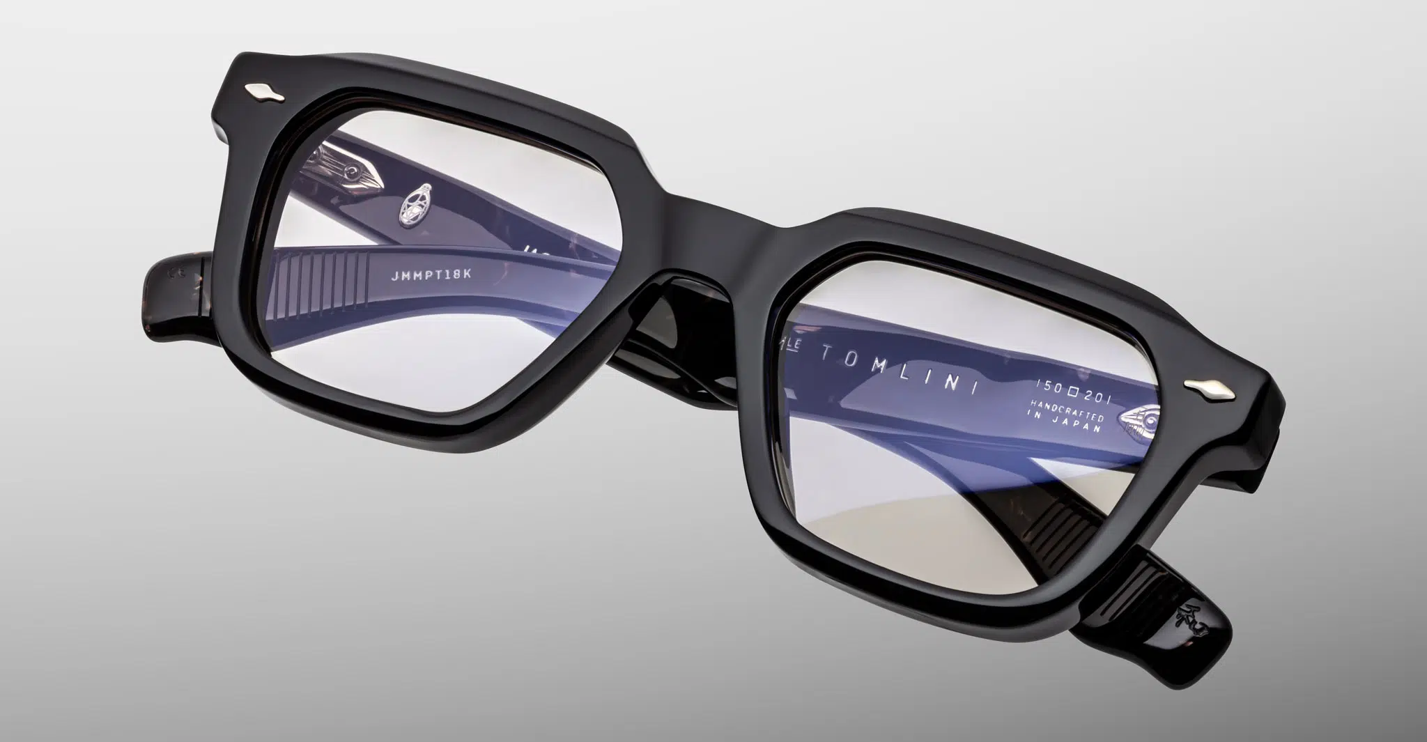 A close-up of black rectangular Jacques Marie Mage Tomlin eyeglasses with thick frames. The inside right temple arm shows the brand name and size. The background is plain light gray.