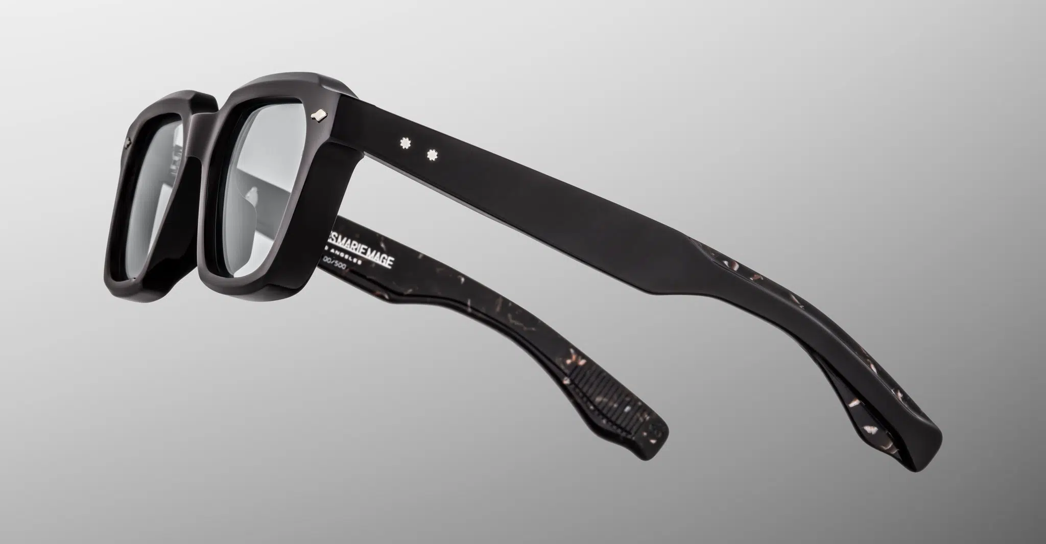 A side view of the Jacques Marie Mage Tomlin eyeglasses showcases their black rectangular thick frames, subtle marbled pattern on the inner temples, and small decorative accents near the hinges, set against a gray background.