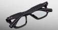 The Jacques Marie Mage Tomlin eyeglasses, featuring thick black frames and clear lenses, are displayed folded against a plain gray background.