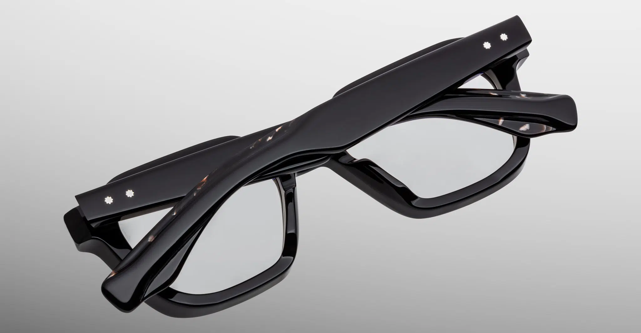 The Jacques Marie Mage Tomlin eyeglasses, featuring thick black frames and clear lenses, are displayed folded against a plain gray background.