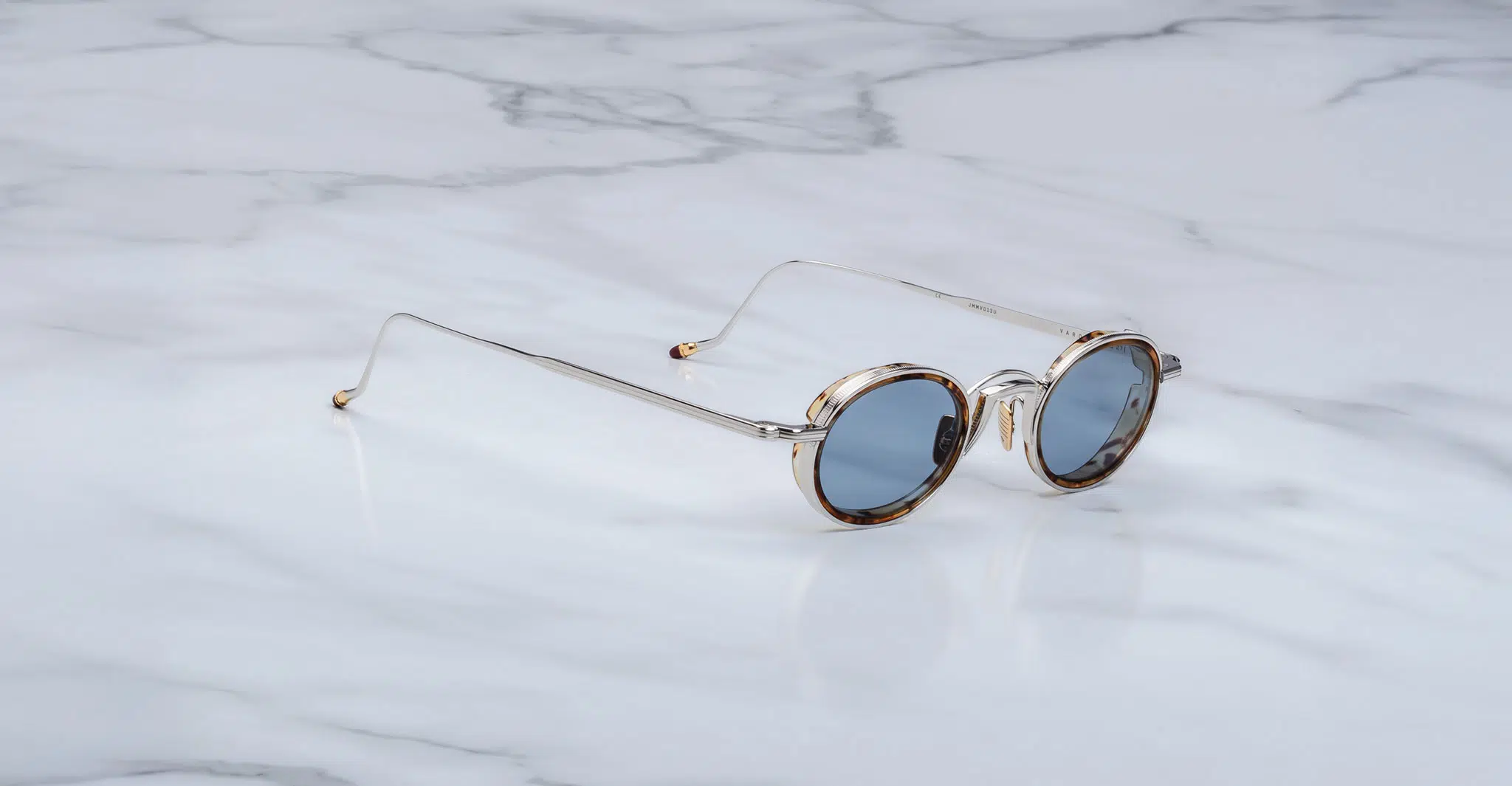 The Jacques Marie Mage Varda round sunglasses with thin gold frames and blue-tinted lenses rest on a white marble surface.