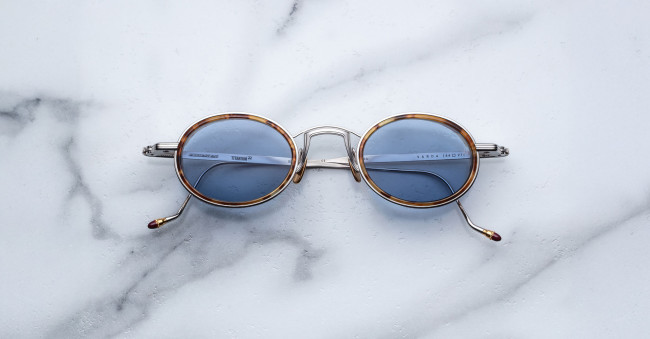 Jacques Marie Mage Varda round sunglasses with thin metal frames and blue-tinted lenses rest on white marble with light grey veining. The arms feature small decorative tips.
