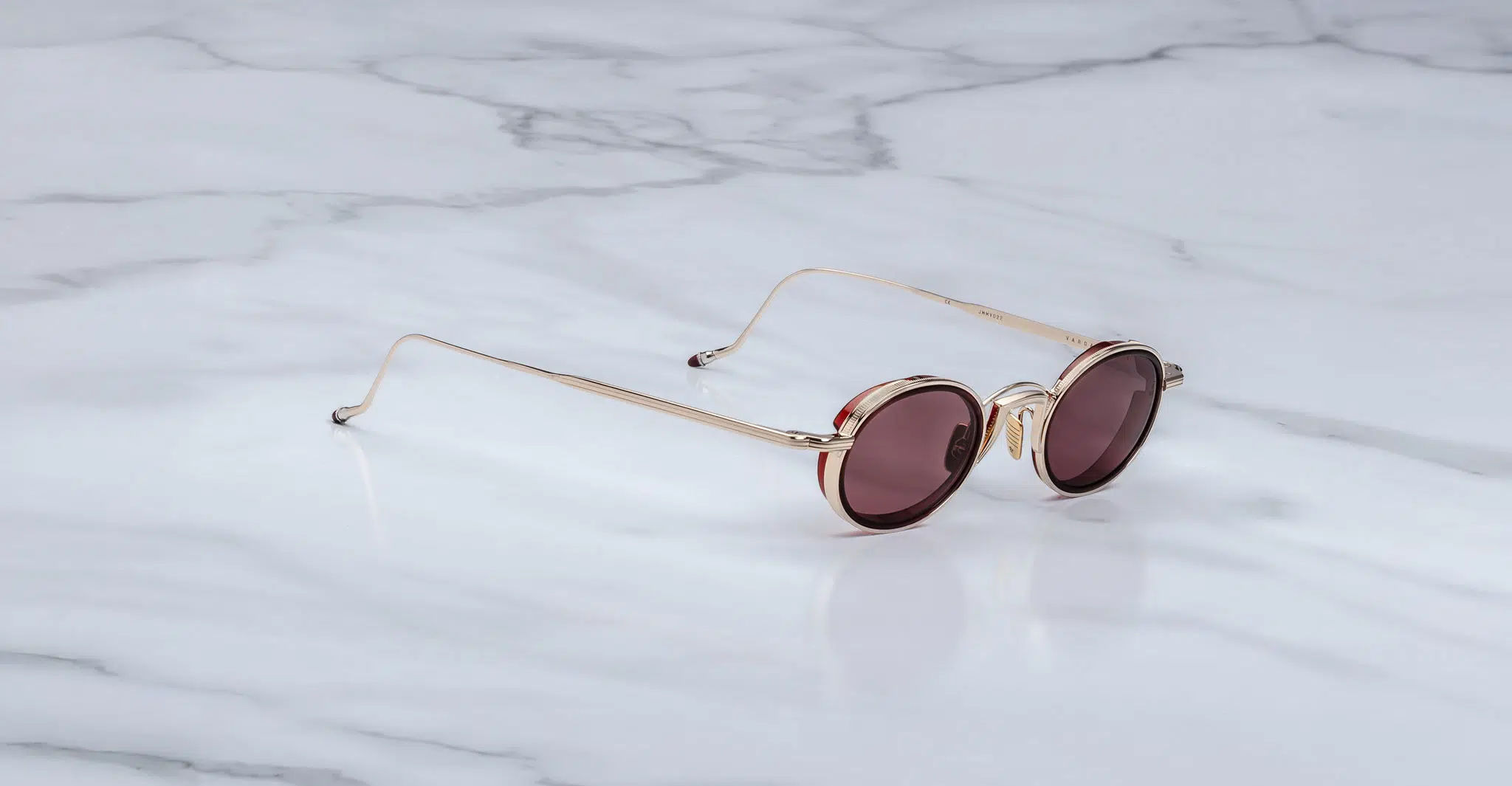 A pair of Jacques Marie Mage Varda round gold-framed sunglasses with dark tinted lenses rests on a white marble surface featuring gray veining.