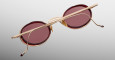 The Jacques Marie Mage Varda features round sunglasses with red-tinted lenses and gold metal frames, displayed from the back with crossed arms against a light grey background.