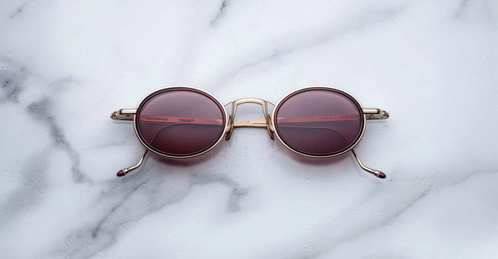 Jacques Marie Mage Varda round sunglasses with gold frames and dark pink tinted lenses rest on a white marble surface, their folded arms and the marbles reflection visible in the lenses.