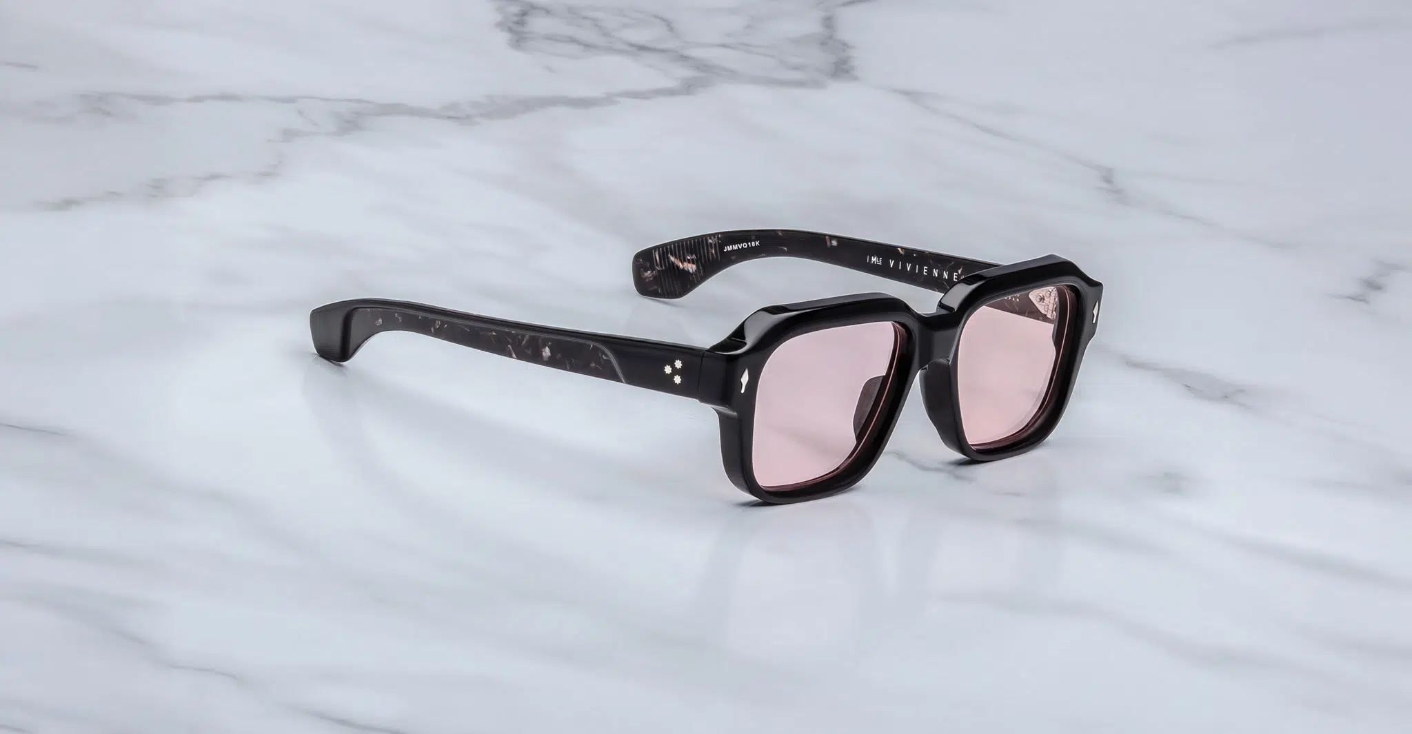 The Jacques Marie Mage Vivienne sunglasses feature a stylish black frame and pink-tinted lenses, elegantly displayed on a white marble surface.