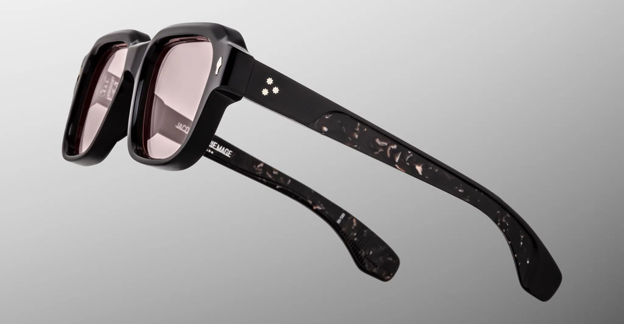 The Jacques Marie Mage Vivienne sunglasses feature thick black frames, slightly tinted lenses, a subtle marbled pattern on the arms, and two small floral accents near the hinges, shown against a light gray background.