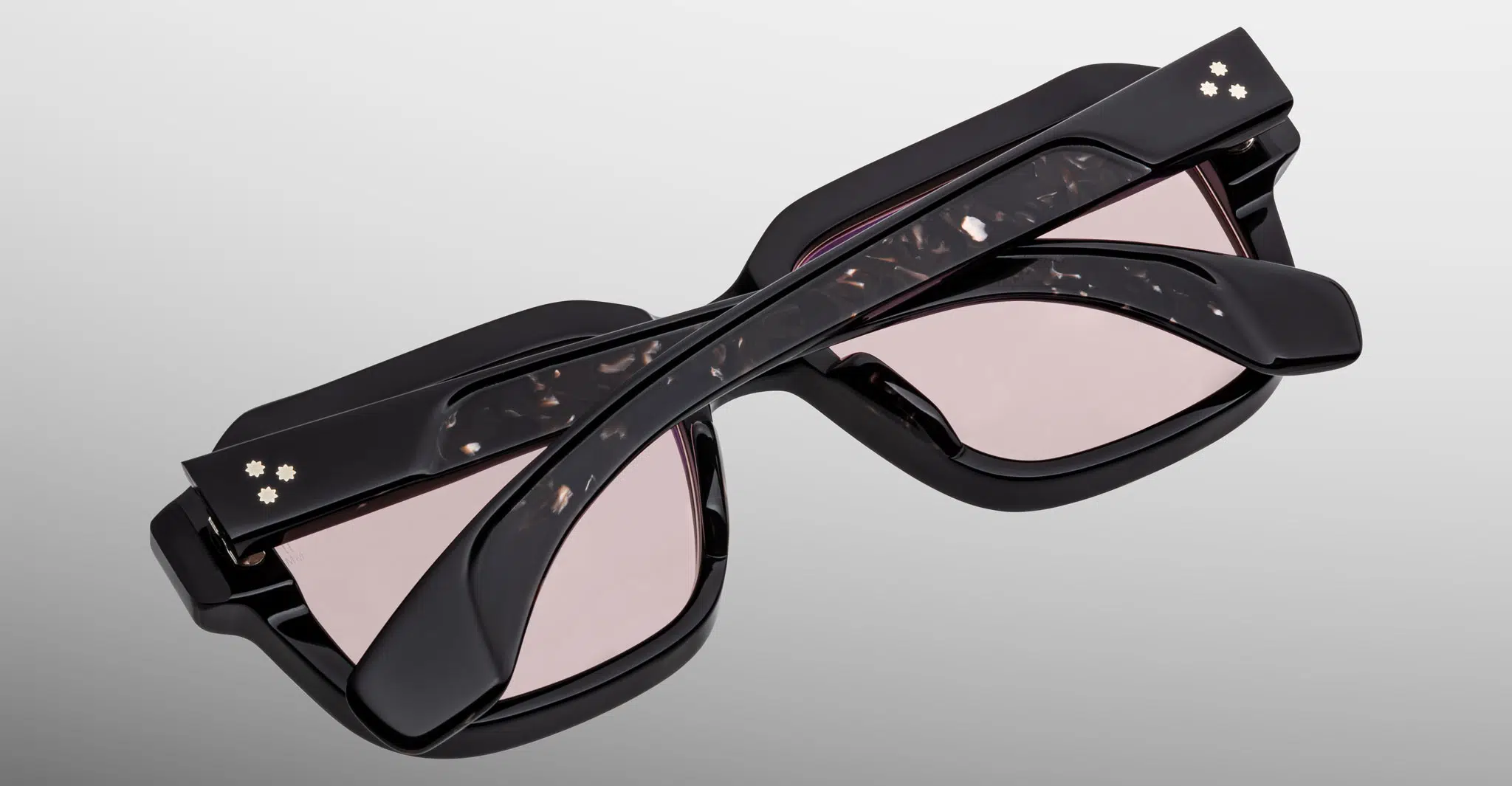 The Jacques Marie Mage Vivienne sunglasses feature a stylish black frame with light pink lenses, marbled temples, and gold accents near the hinges. Shown folded from above on a plain gradient light gray background.