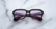 The Jacques Marie Mage Vivienne is a pair of black rectangular sunglasses with purple-tinted lenses, shown resting on a white marble surface.