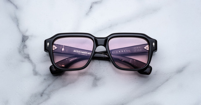 The Jacques Marie Mage Vivienne is a pair of black rectangular sunglasses with purple-tinted lenses, shown resting on a white marble surface.