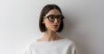 A person with a straight dark bob wears oversized green Lapima Alice glasses, standing against a plain white wall.