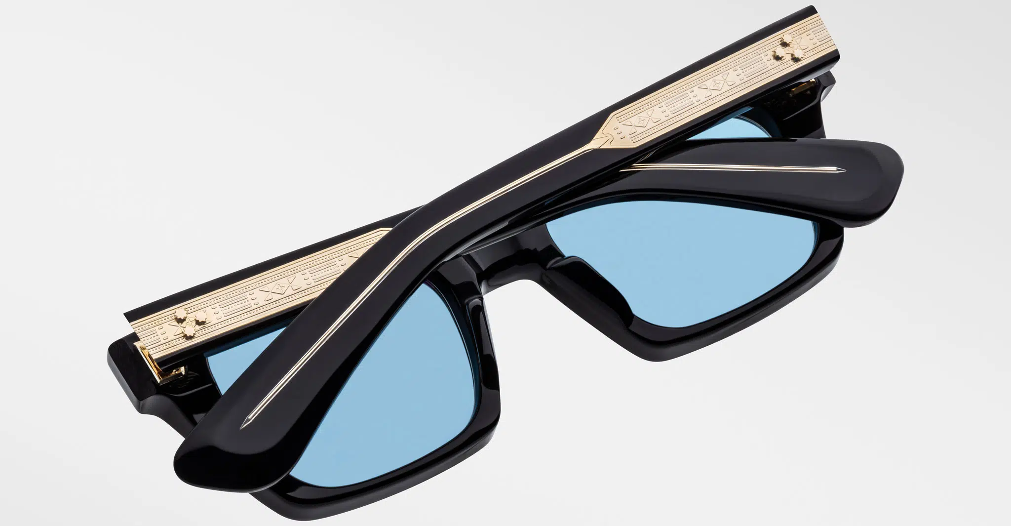 A pair of black sunglasses with blue lenses, shown folded. The frame features a gold metal accent with engraved detailing along the top edge. The background is plain light gray.