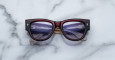 A pair of Jacques Marie Mage Chelsea cherry red, thick-framed sunglasses with light purple lenses rests on a white marble surface with grey veins, with faint brand details visible on the inside of the left lens.