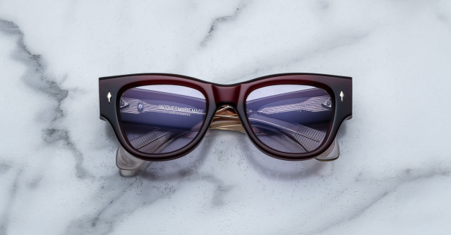 A pair of Jacques Marie Mage Chelsea cherry red, thick-framed sunglasses with light purple lenses rests on a white marble surface with grey veins, with faint brand details visible on the inside of the left lens.